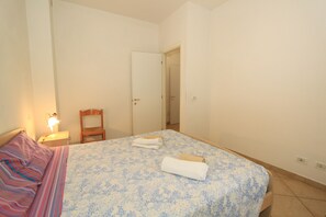 A bedroom with a bed, a nightstand, a chair, and a door leading to another room. - San Remo Apartments (Comacchio)