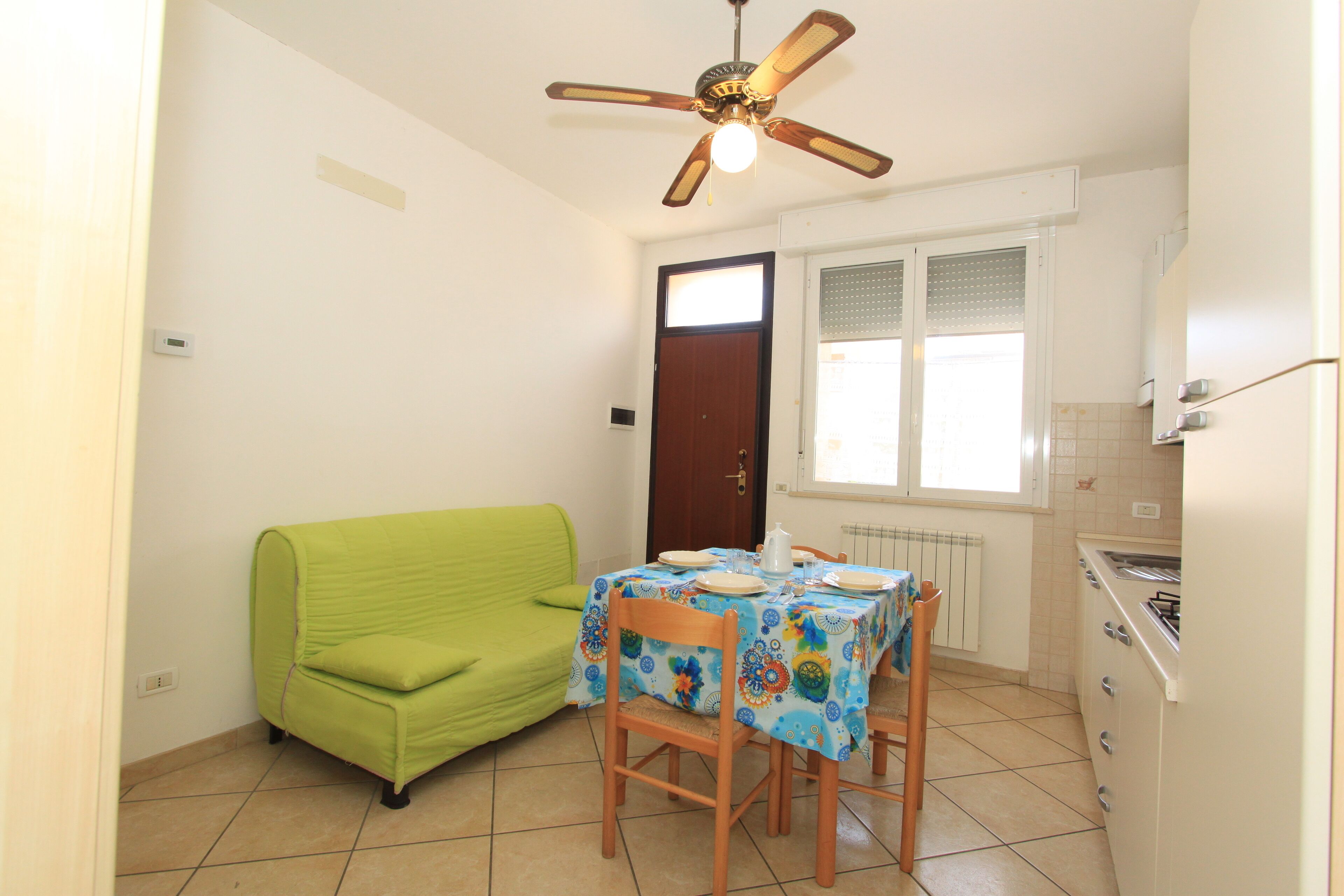 A compact kitchen with a dining table, chairs, a green sofa, a ceiling fan, and a window with blinds.