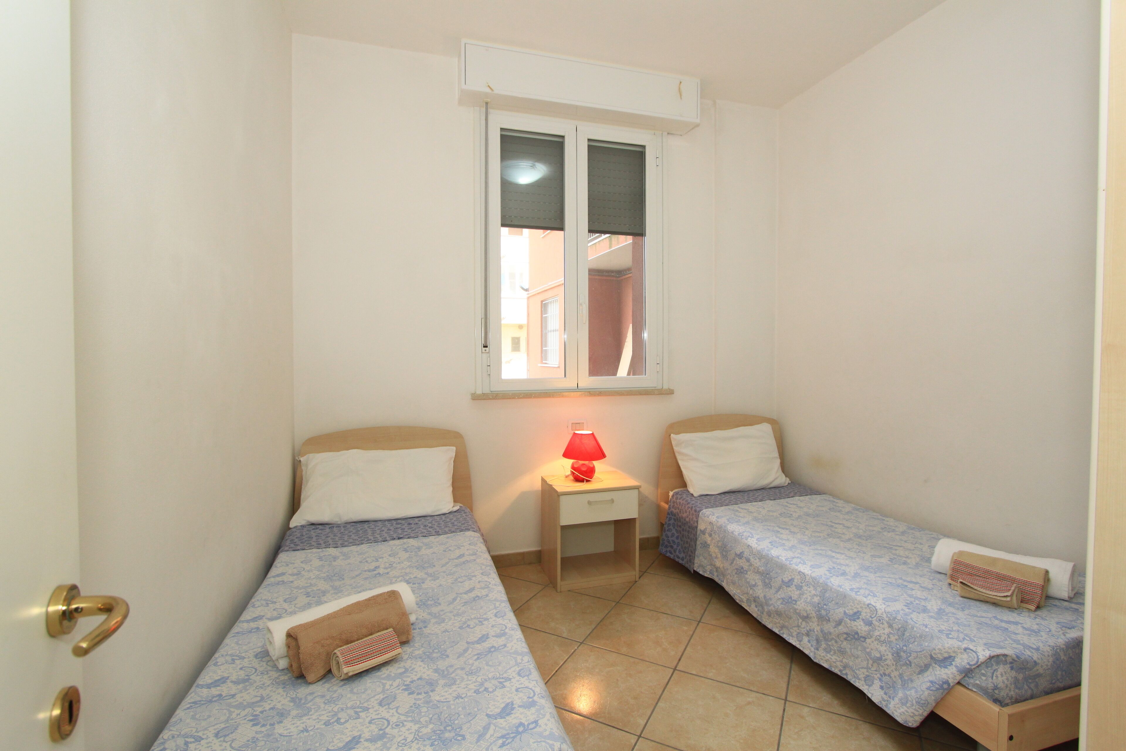 A small, clean room with two single beds, a nightstand with a lamp, and a window with blinds.