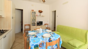 Un comedor con una mesa dispuesta para cuatro personas, un sofá verde, un ventilador de techo y un televisor sobre una estantería.