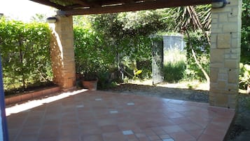 Uma varanda coberta com piso de azulejos, pilares de pedra e vista para um jardim com diversas plantas e um portão.