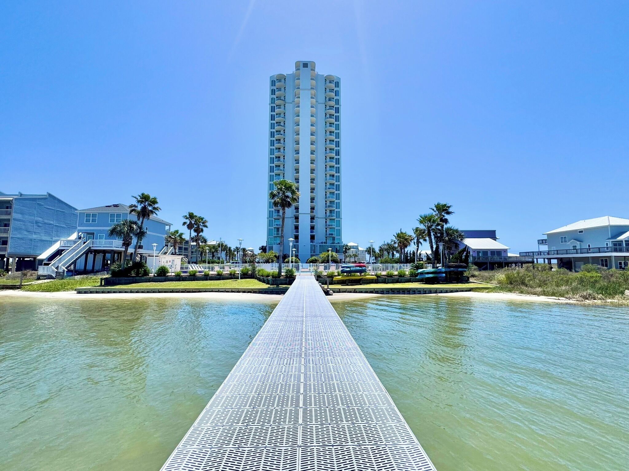 A waterfront hotel with a central walkway leading to a tall building, surrounded by palm trees and a clear sky.