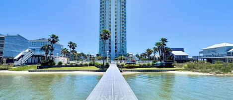 A waterfront hotel with a central walkway leading to a tall building, surrounded by palm trees and a clear sky.