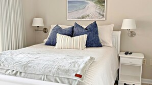 A neatly made bed with a white comforter and pillows, a bedside table with a lamp, and a framed beach scene on the wall.