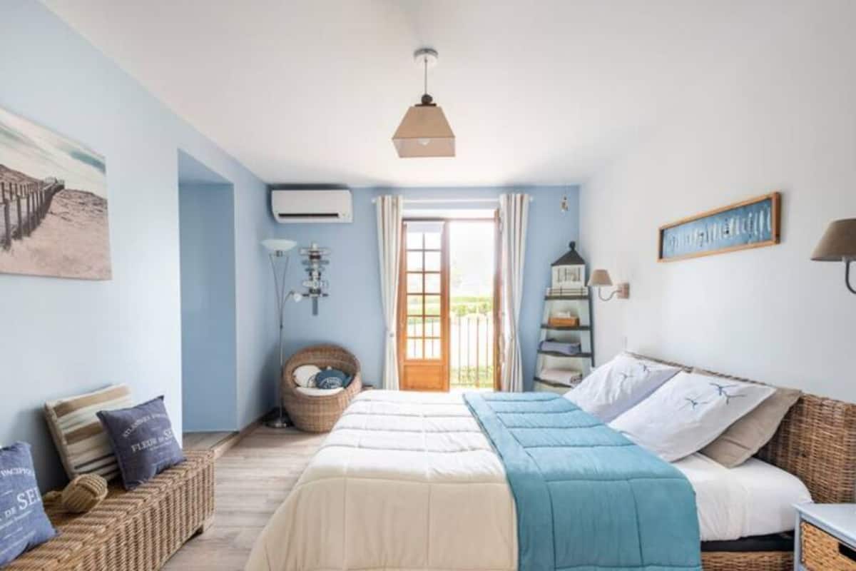 A bedroom with a large bed, a bedside table, a lamp, a wicker chair, and a view of the outdoors through a door.