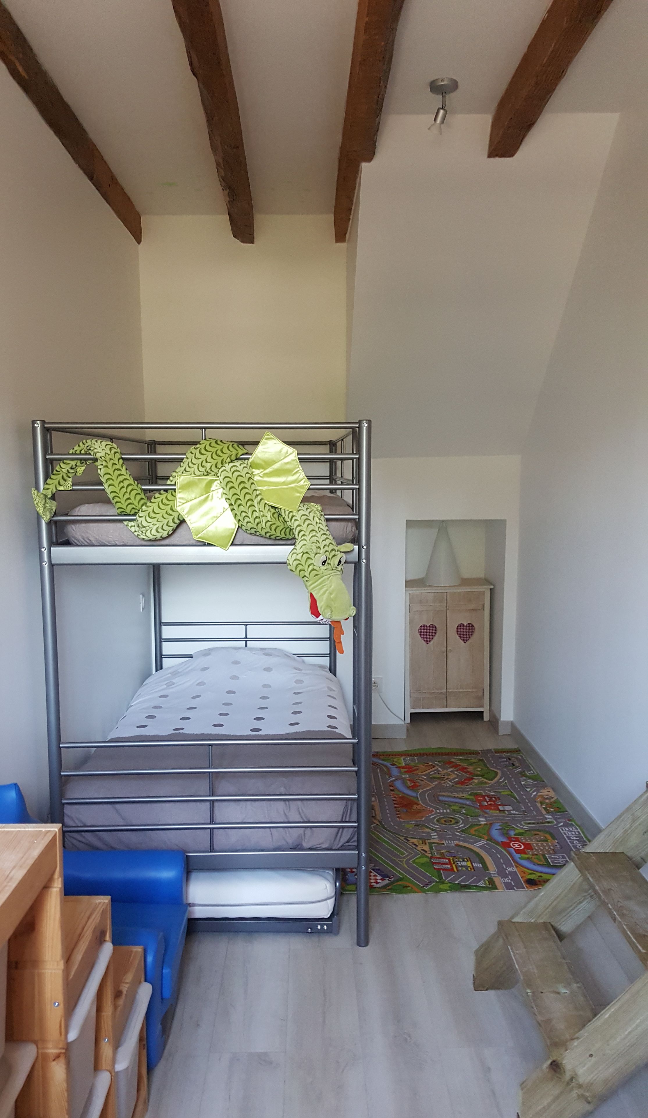 A bunk bed with a green dinosaur decoration, a wooden cabinet with heart designs, and a colorful play mat on the floor.