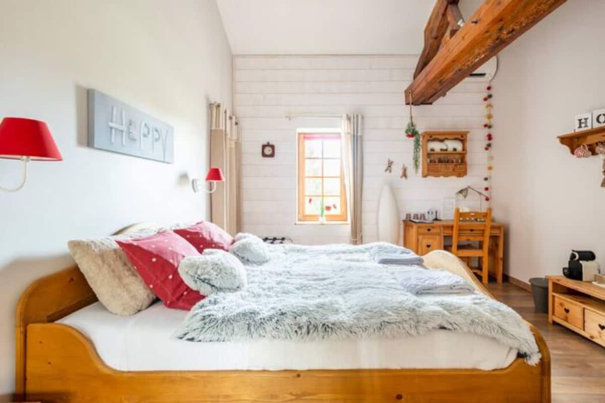 A cozy bedroom with a wooden bed, a desk, and a chair. There is a window with curtains, a wall-mounted lamp, and a shelf with decorations.