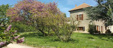 A rustic house with a stone facade, a tiled roof, and a garden with blooming trees.