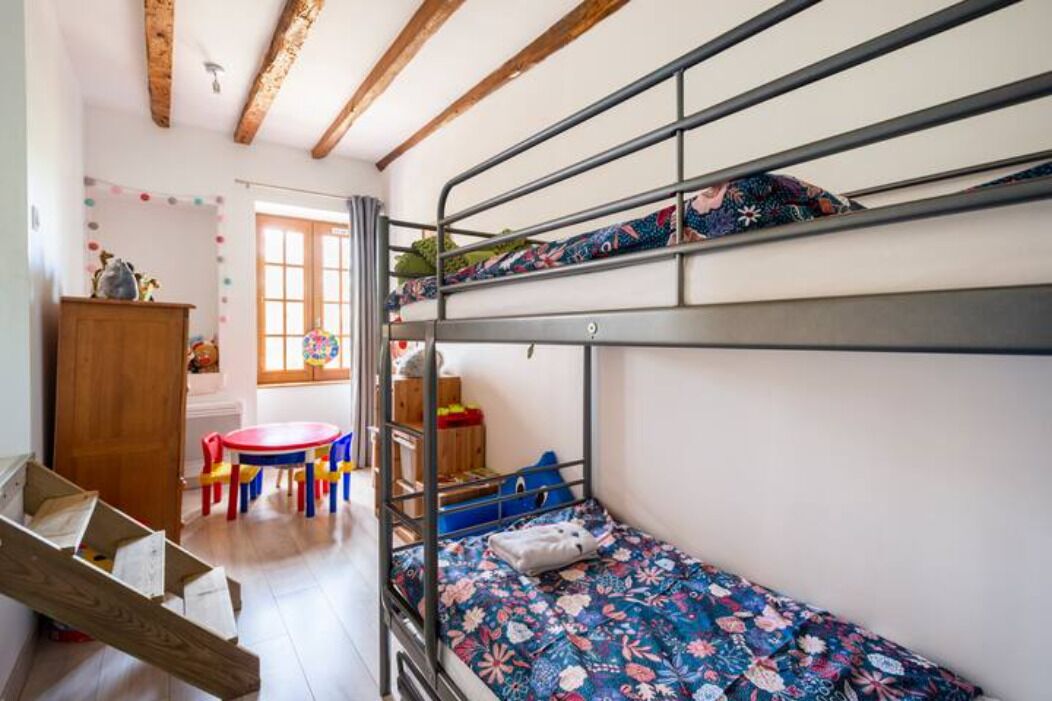 A bunk bed with a floral bedspread, a wooden table with chairs, and a window with curtains.