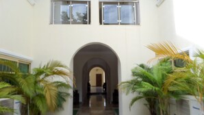 A corridor with an arched entrance, flanked by potted palm trees, leading to a room with a bed and a window. - Nordic Hospitality Hotel (Abuja)
