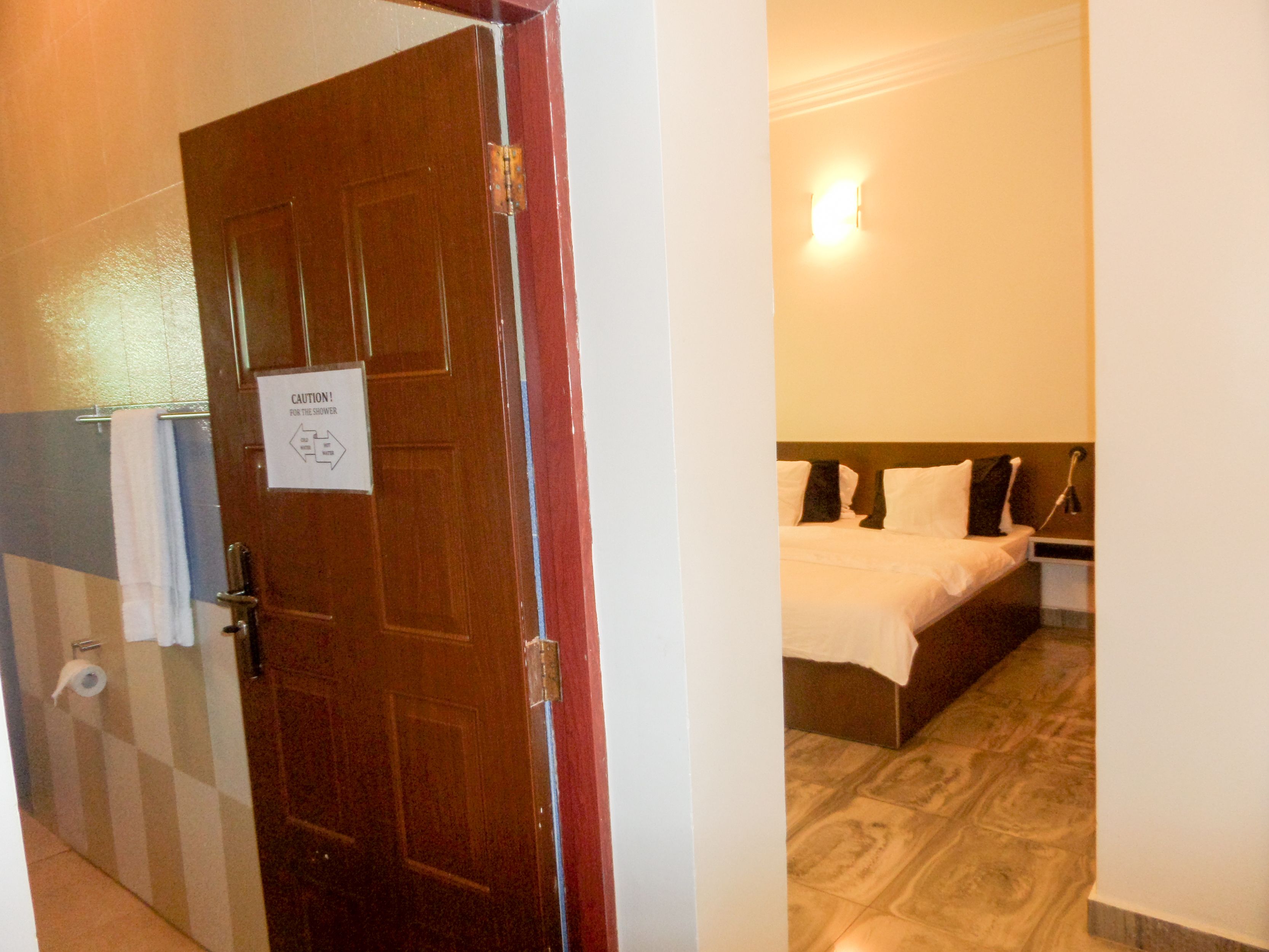 A hotel room with a wooden door, a sign indicating caution for the shower, a bed with white linens, and a wall-mounted light fixture.