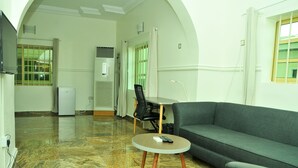 A modern living room with a grey sofa, a round coffee table, and a black chair. There is a flat-screen TV mounted on the wall, a small refrigerator, and a window with bars. - Nordic Hospitality Hotel (Abuja)