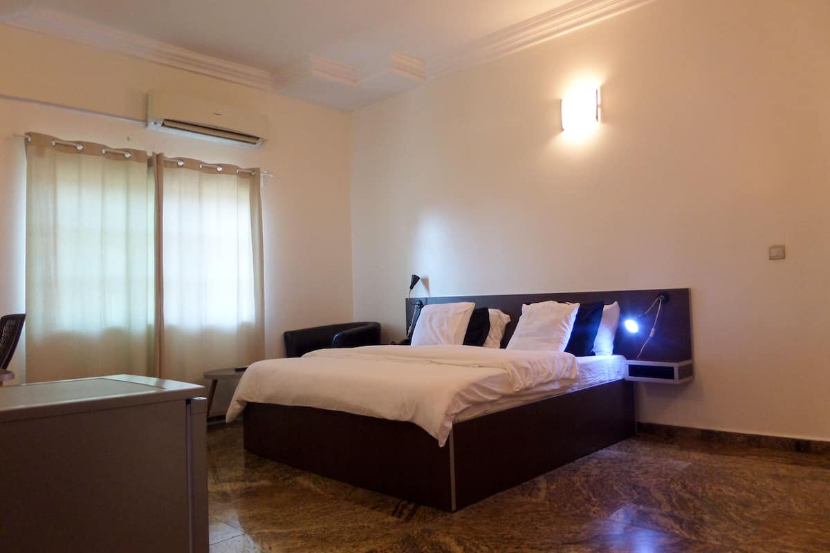 A hotel room with a bed, a desk, and a chair. There is a window with curtains, a wall-mounted light, and an air conditioning unit.