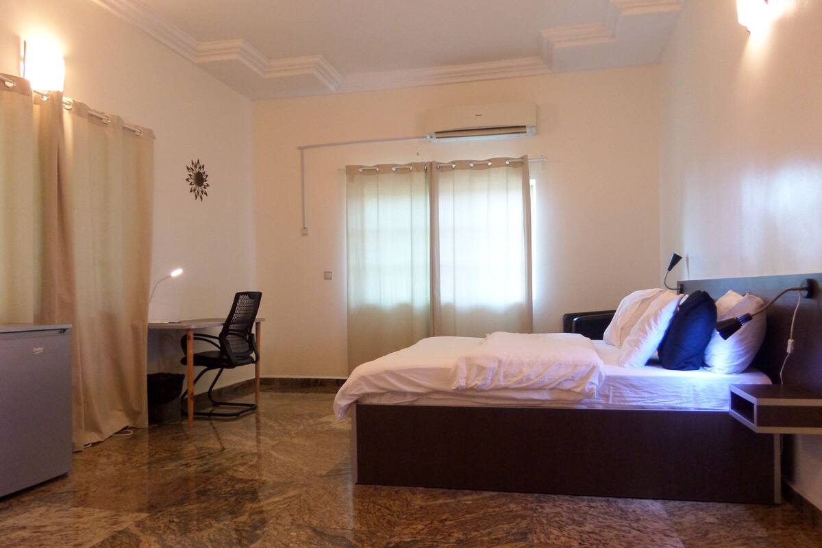 A hotel room with a bed, a desk, a chair, and an air conditioning unit.
