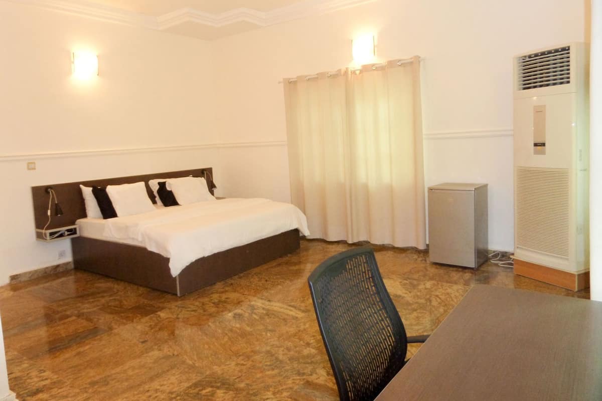 A hotel room with a bed, a desk, a chair, a window with curtains, and an air conditioning unit.