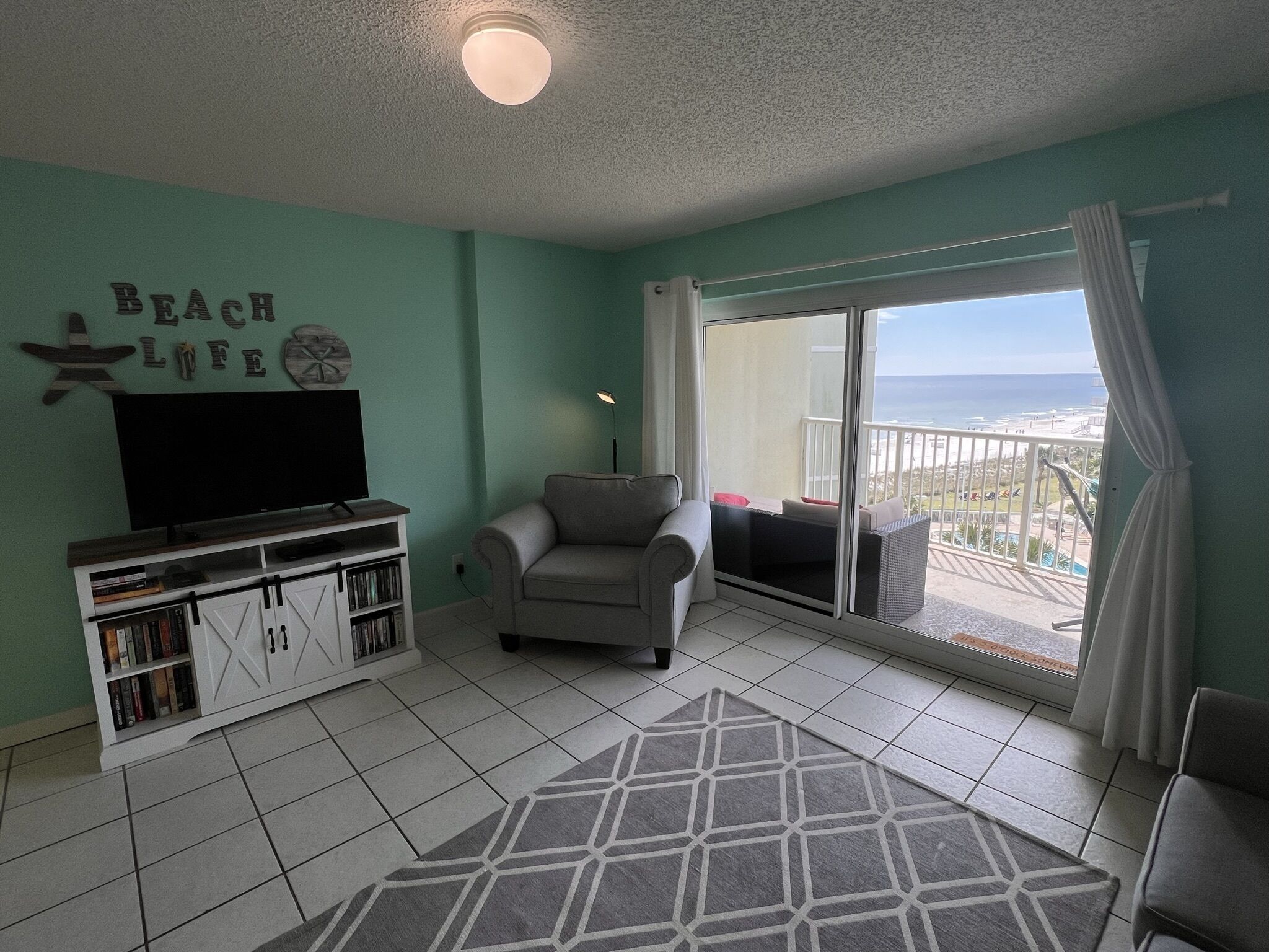 A living room with a flat-screen TV, a grey sofa, a bookshelf, and a view of the ocean through a sliding glass door.