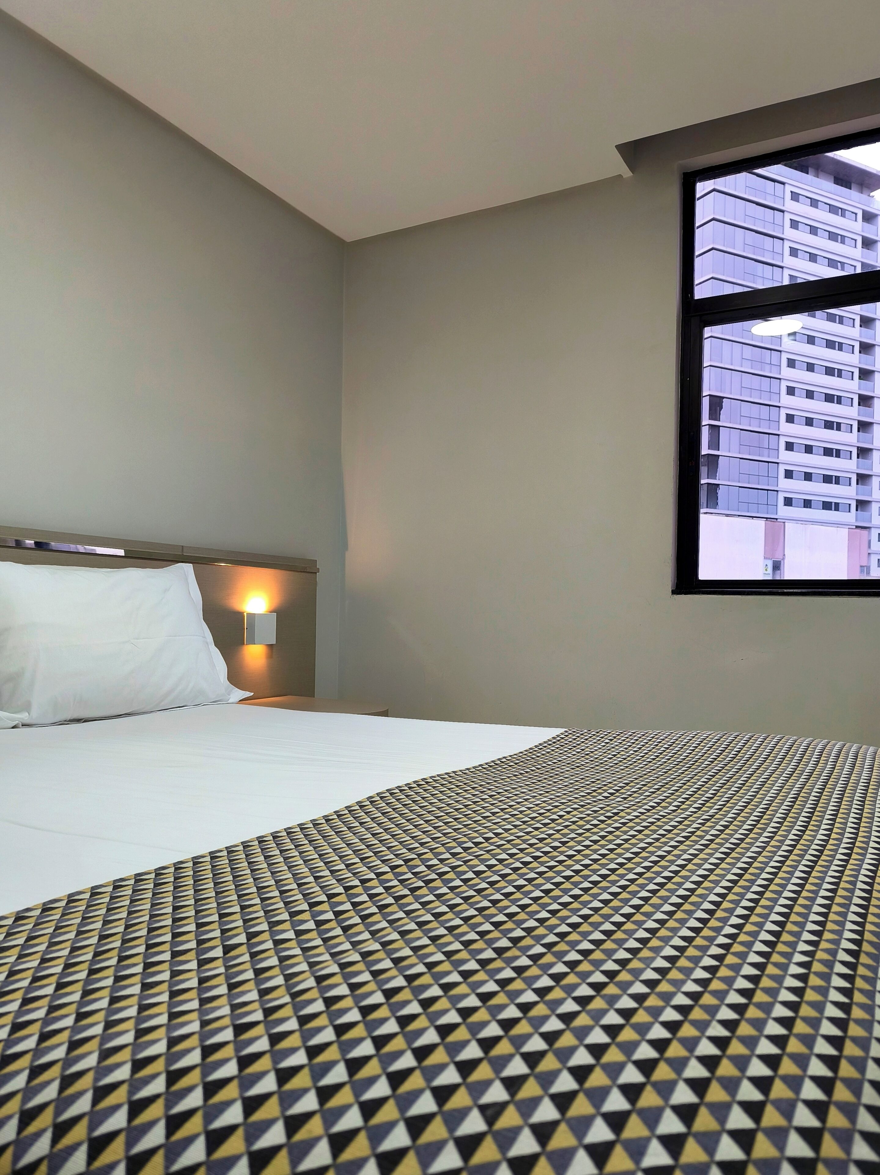 A hotel room with a bed, a nightstand, a wall-mounted lamp, and a window with a view of a building.