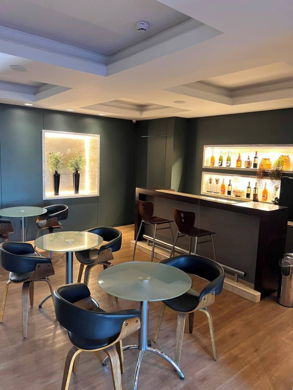 Un área de bar moderna con mesas y sillas redondas, una barra con botellas y una ventana con plantas.