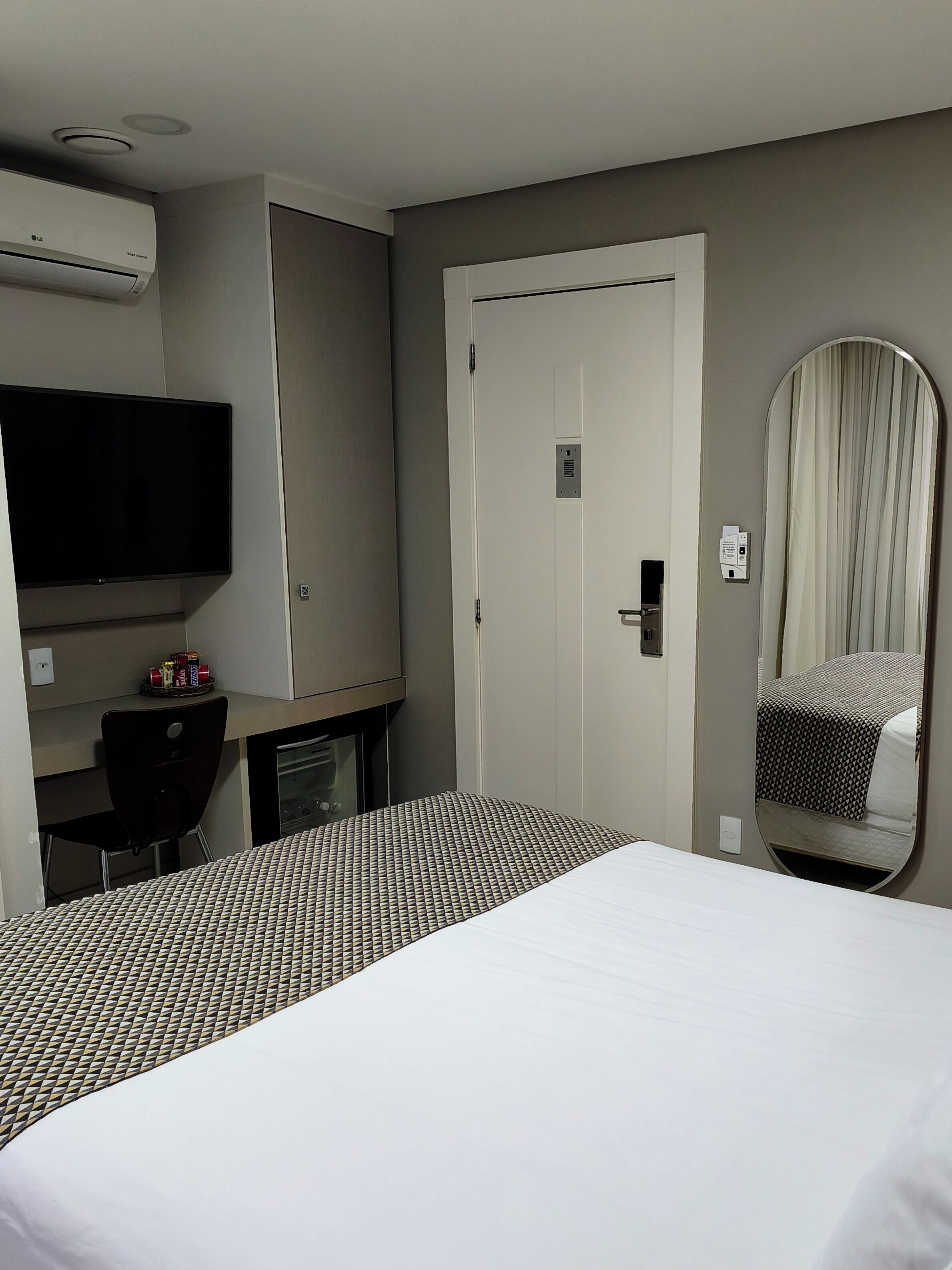 Habitación de hotel con cama, escritorio, televisor, espejo y aire acondicionado.