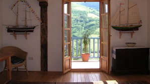 A room with a wooden ceiling, a wooden chair, a table, a potted plant, and a view of a green landscape through an open door.