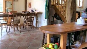 A rustic kitchen with wooden beams, a dining table with chairs, and a fruit bowl on a wooden stand.
