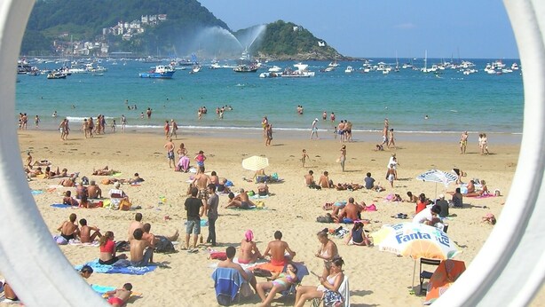 A beach scene with a clear view of the sea, boats, and a hill with buildings.