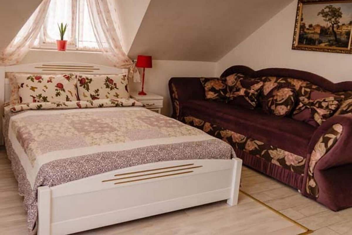 Ein Schlafzimmer mit Bett, einer Couch, einem Nachttisch, einer Lampe und einem Fenster mit Pflanze.