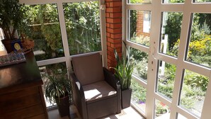 Een zonnige kamer met een rieten bank, potplanten en uitzicht op groen.