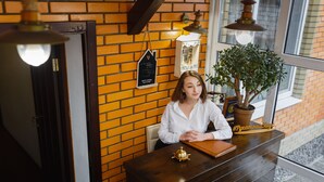 Een vrouw zit achter een houten receptiebalie in een ruimte met bakstenen muren, een kamerplant en een raam met uitzicht.
