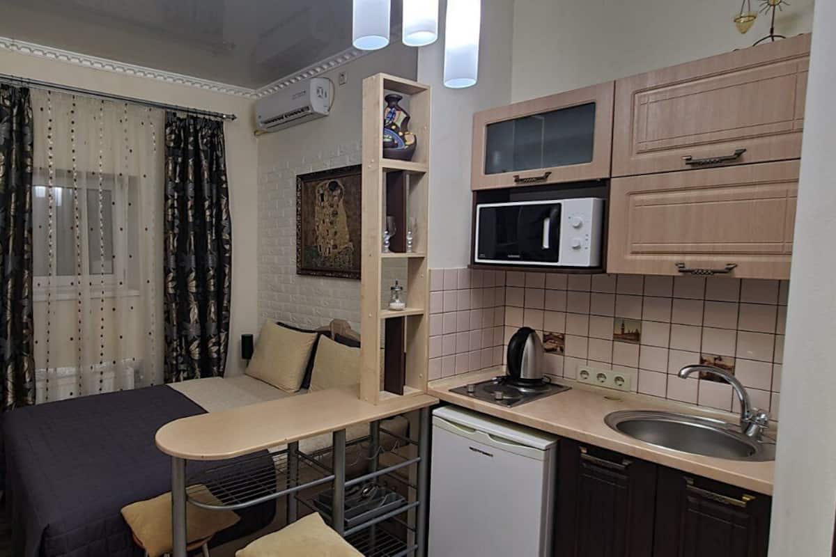 Ein kleines Zimmer mit Bett, Schreibtisch, Mikrowelle, Spüle und einer kompakten Küchenzeile.