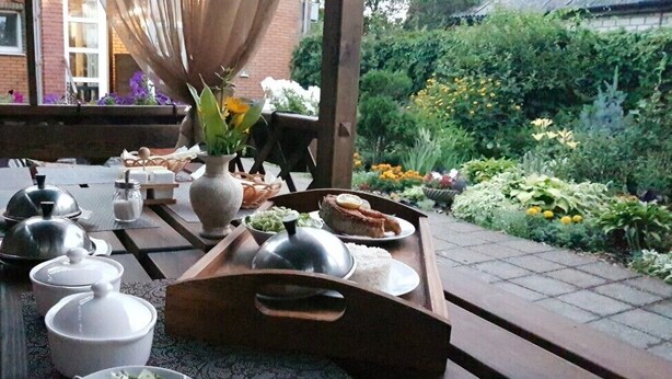 Een buitenterras met een houten tafel, gedekt voor een maaltijd, omringd door een tuin met diverse planten en bloemen.