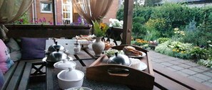 Un área de comedor al aire libre con una mesa de madera dispuesta para una comida, rodeada por un jardín con diversas plantas y flores.