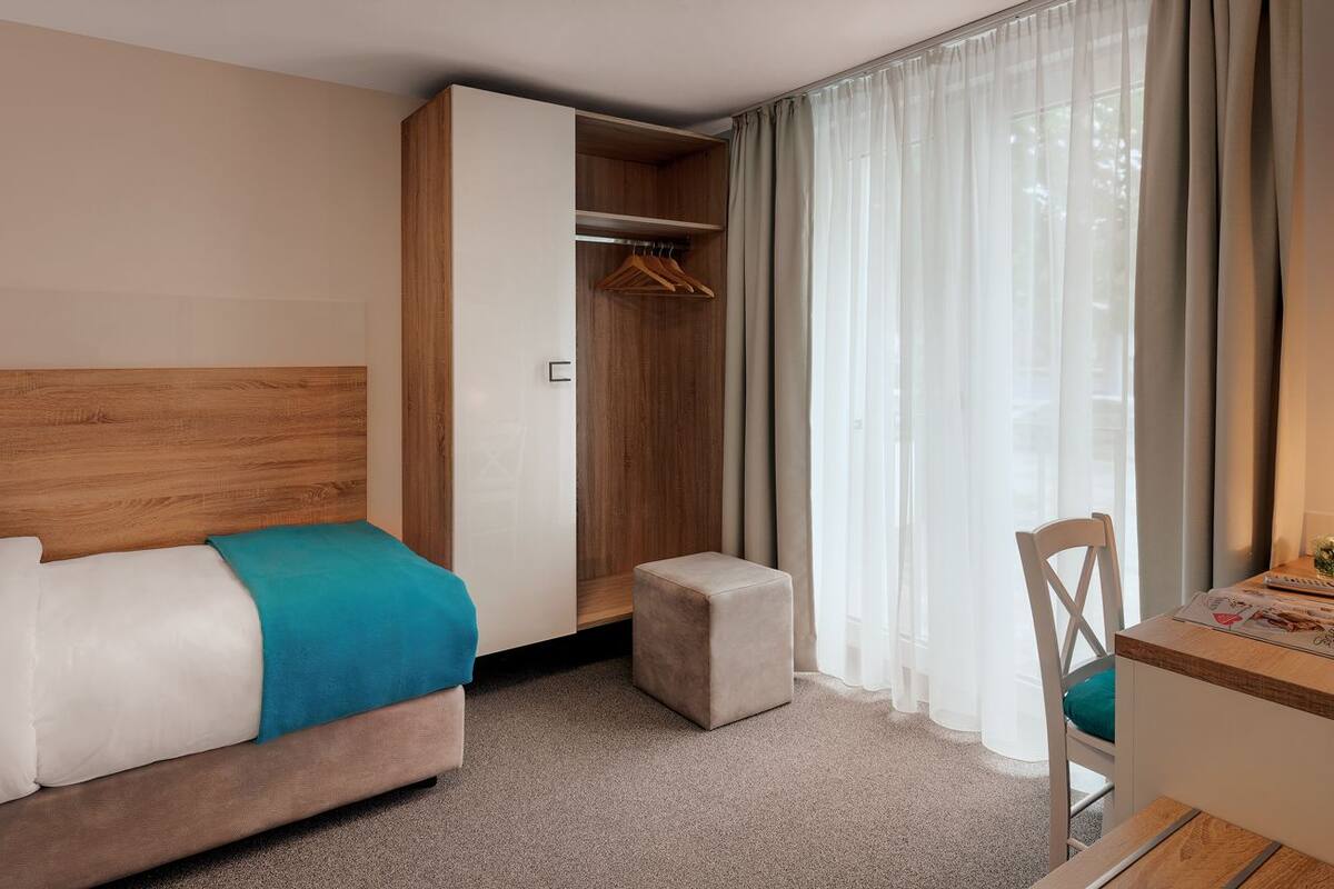 A hotel room with a bed, a wooden headboard, a closet with hanging clothes, a small table with a chair, and a window with curtains.