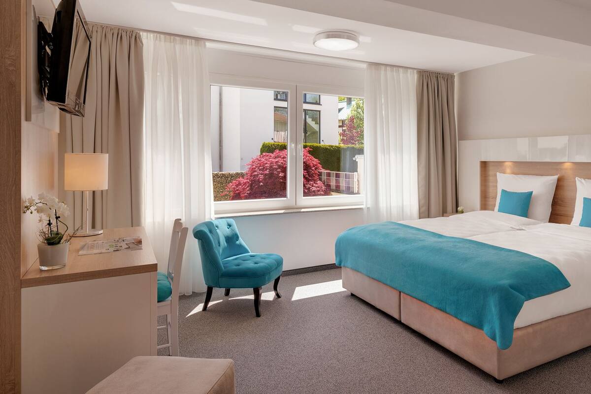 A modern hotel room with a large bed, a desk with a lamp, a chair, and a window with curtains.