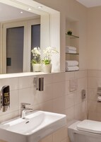 Un baño moderno con un espejo grande, lavabo blanco y retrete. Hay dos orquídeas en maceta sobre el estante del espejo.