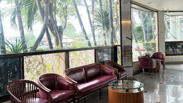 Un vestíbulo amplio con un sofá de cuero, sillas de madera y una mesa de centro de vidrio. Amplias ventanas ofrecen vistas a palmeras y un edificio en el exterior.