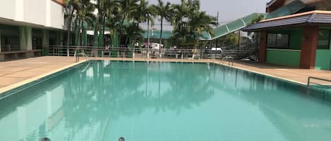 A swimming pool with clear blue water, surrounded by palm trees and a building with a green and white facade.