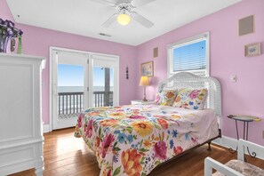 A bedroom with a floral bedspread, a white ceiling fan, a window with blinds, and a view of the outdoors.