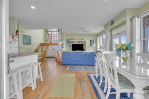 A spacious living area with a blue sofa, white chairs, a glass-top dining table, and a staircase leading to an upper level.