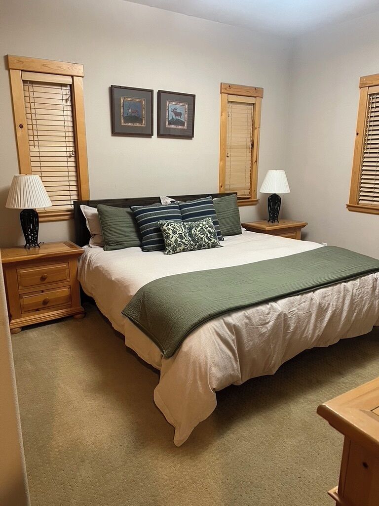 Un dormitorio con cama, dos mesitas de noche, una lámpara en cada mesita y una puerta de madera, además de dos cuadros en la pared.