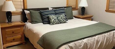 Un dormitorio con cama, dos mesitas de noche, una lámpara en cada mesita y una puerta de madera, además de dos cuadros en la pared.