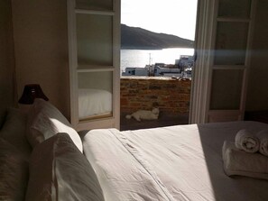 A bedroom with a bed, a window with a view of a body of water and buildings, and a cat on the balcony. - Diosmarini Studios & Apartments (Amorgos)