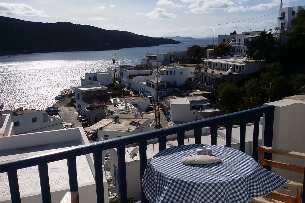 A balcony with a table and chairs overlooking a coastal town with white buildings and a view of the sea.