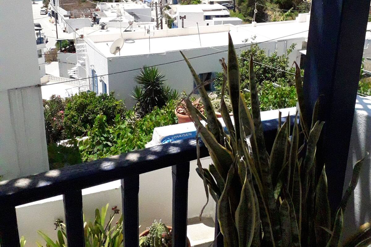 A balcony with potted plants, overlooking a coastal town with white buildings and a view of the sea.