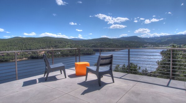 Un balcon avec deux chaises et un petit tabouret orange, donnant sur un lac et des montagnes boisées.