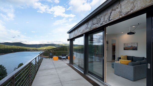 Een modern balkon met glazen hekwerk met uitzicht op een meer, een bank en een schilderij aan de muur.
