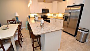 A modern kitchen with white cabinets, a stainless steel refrigerator, a granite countertop, and a dining area with wooden chairs and a table. - Waves, Sand, and Fun in the Sun come join us for a great time. 🌞🍂 (Gulf Shores)
