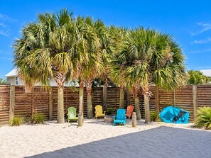 A sandy beach area with palm trees, a wooden fence, and colorful Adirondack chairs. - A Beach Life 3A | 3BR Beach Getaway + Pool (Gulf Shores)