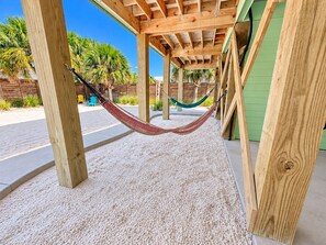 A hammock under a wooden pergola with a sandy ground, surrounded by greenery and a green building. - A Beach Life 3A | 3BR Beach Getaway + Pool (Gulf Shores)