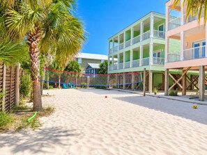 A beach volleyball court with a net, palm trees, and a multi-story building with balconies. - A Beach Life 3A | 3BR Beach Getaway + Pool (Gulf Shores)